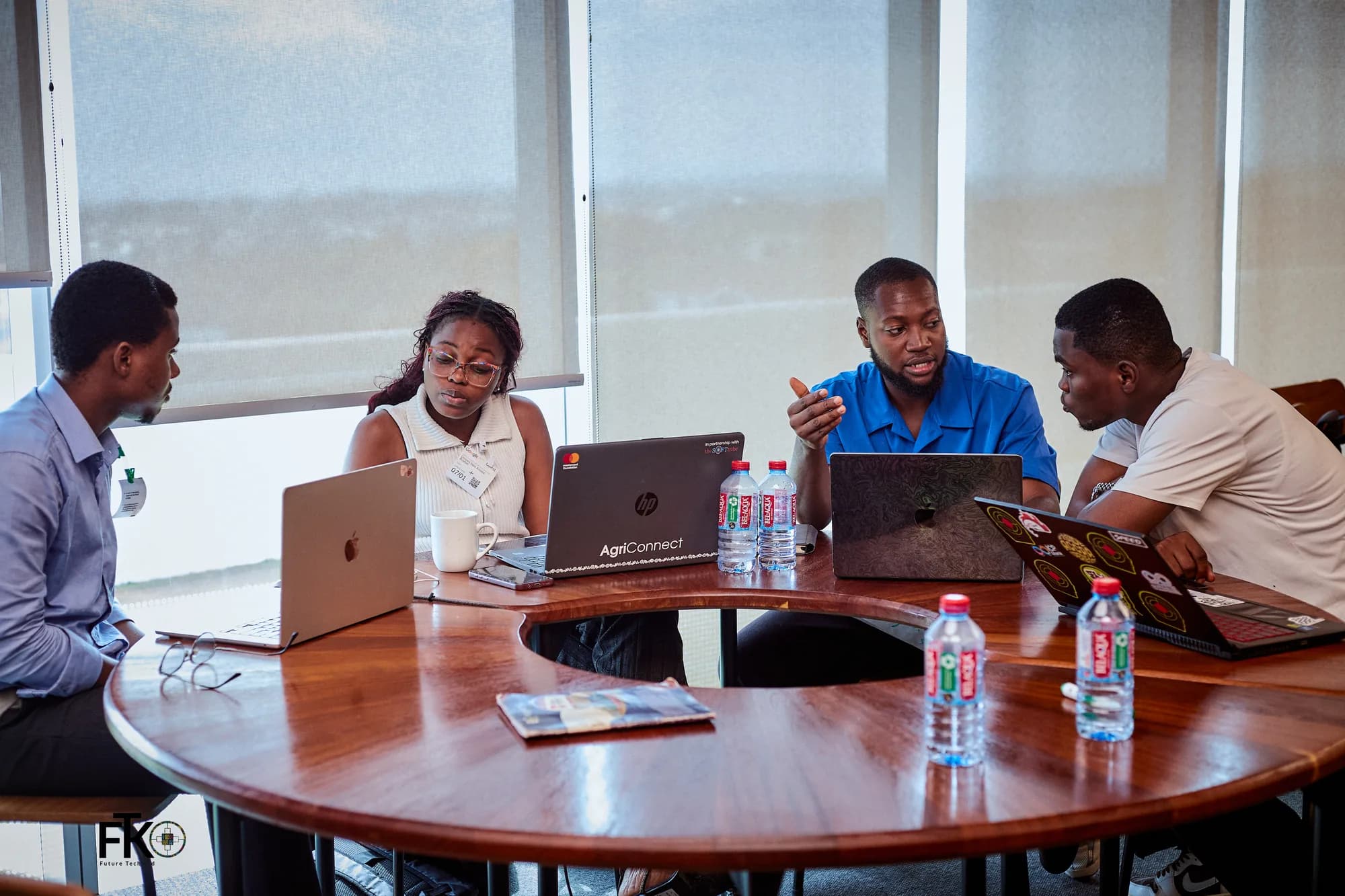 A team of four at their laptops around a curved table, one of them talking an idea through