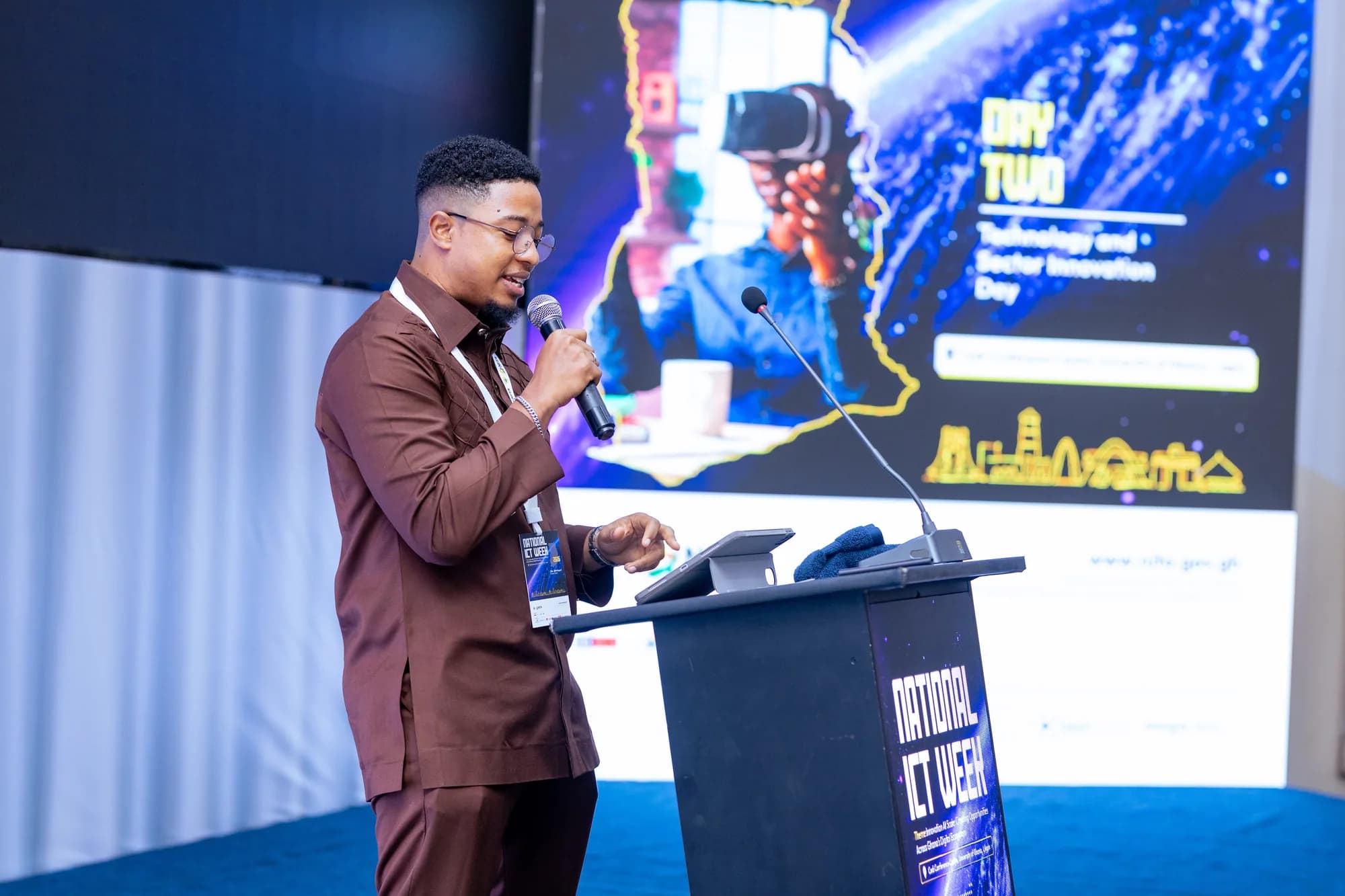 Toby Woode speaking at the National ICT Week podium, in front of the Day Two, Technology and Sector Innovation Day board